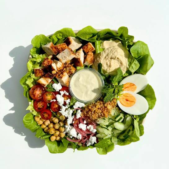 Salada de frango, hummus, queijo de cabra, grão-de-bico, tomate grape, pepino, ovo, cebola, crocante de quinoa, hortelã, folhas crocantes, molho iogurte-tahine